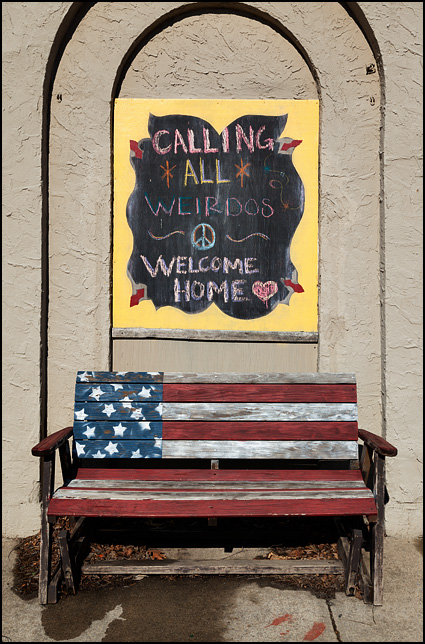 An old wooden glider bench painted like the American flag under a sign that says, Calling All Weirdos Welcome Home. It sits next to Little Shop of Lauras on Broadway in Fort Wayne, Indiana.