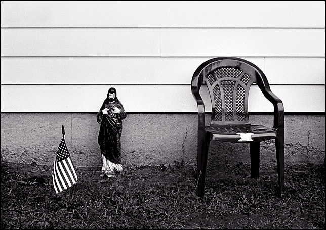 A small American flag and a statue of Jesus Christ stand next to a plastic patio chair in front of a house on Wells Street in Fort Wayne, Indiana.